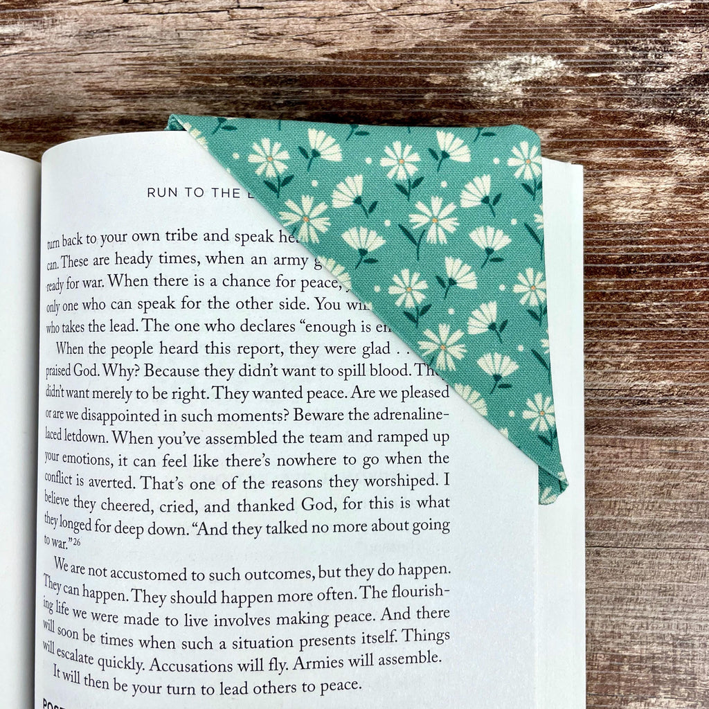 May include: A teal fabric bookmark with a white floral pattern is placed on the corner of a book. The bookmark is triangular and folded over the pages. The book's pages are visible, with text from the book visible.