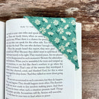 May include: A teal fabric bookmark with a white floral pattern is placed on the corner of a book. The bookmark is triangular and folded over the pages. The book's pages are visible, with text from the book visible.
