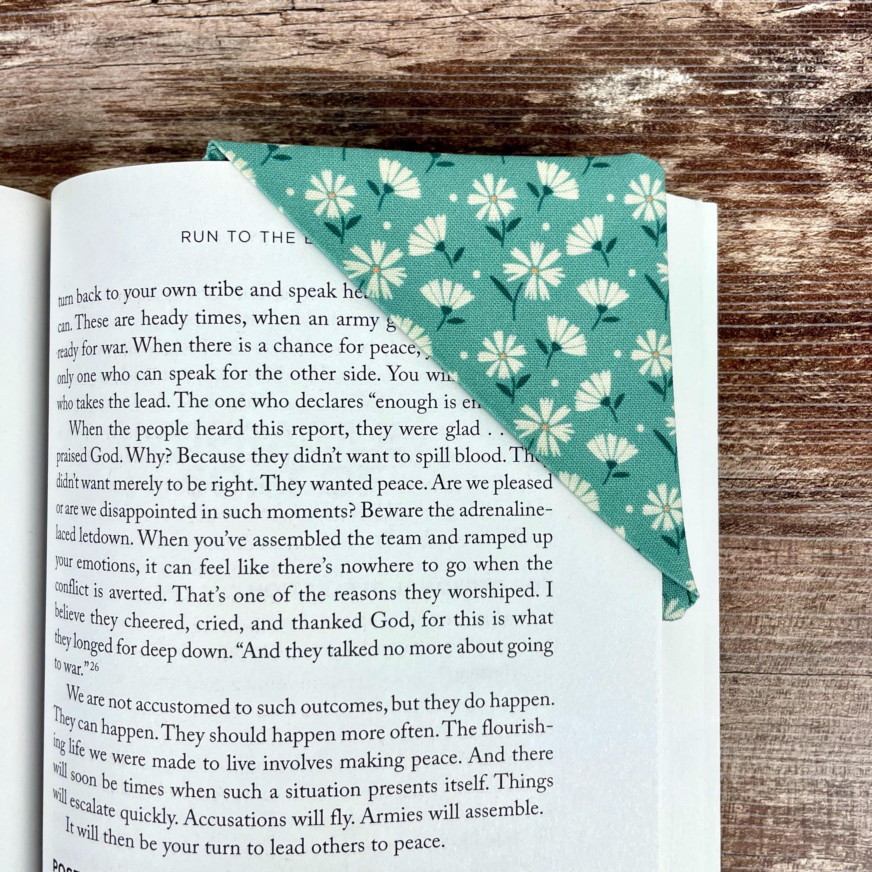 May include: A teal fabric bookmark with a white floral pattern is placed on the corner of a book. The bookmark is triangular and folded over the pages. The book's pages are visible, with text from the book visible.